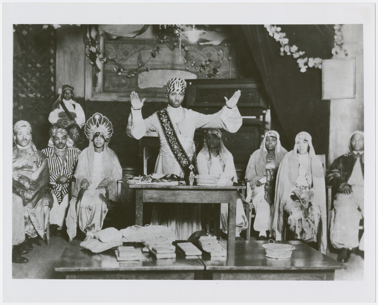 Image of Noble Drew Ali standing at a table, while several temple members are seated behind him.